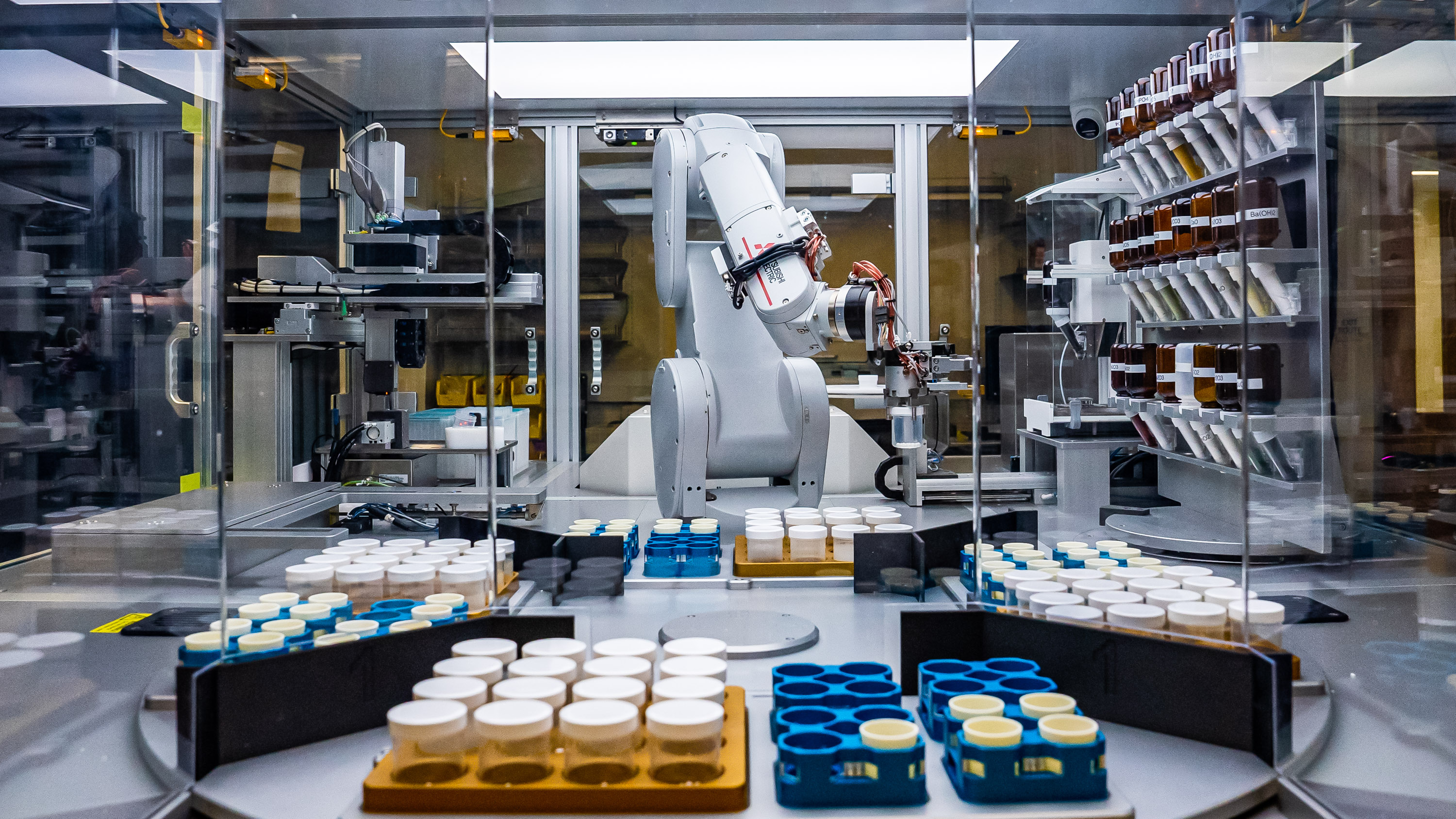 a robotic arm in a plexiglass-enclosed workspace filled with sample bottles