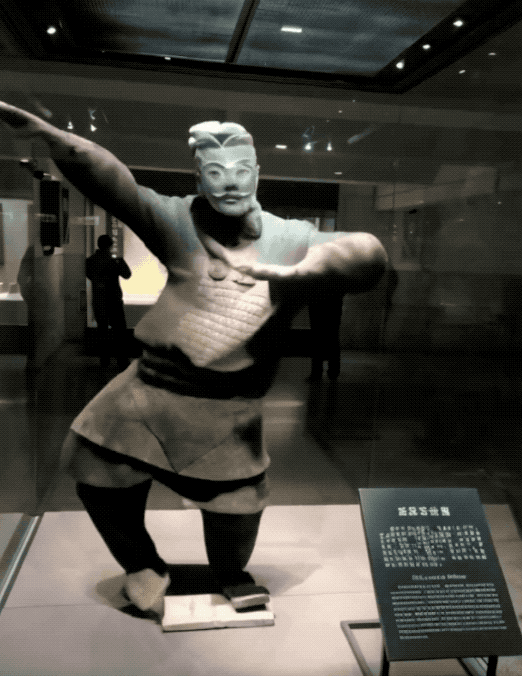 A terra-cotta warrior in a museum, doing an expressive dab pose as part of a viral dance routine.